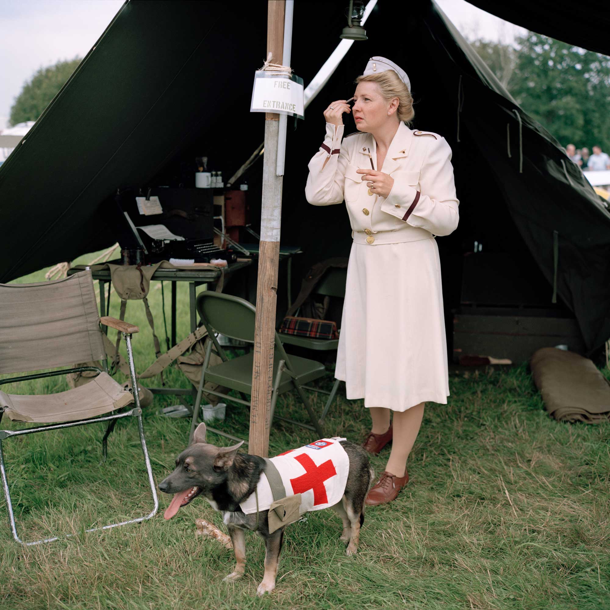 A woman dressed as a World War II-era nurse applies makeup outside a military-style tent.