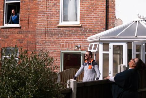 &quot;PICTURING LOCKDOWN&quot;, A GARDEN SCENE ON A SUNNY DAY WITH BLUE SKY, TWO PEOPLE LAUGING OVER GARDEN FENCES AS ANOTHER LOOKS DOWN FROM A FIRST FLOOR WINDOW ALSO LAUGHING.