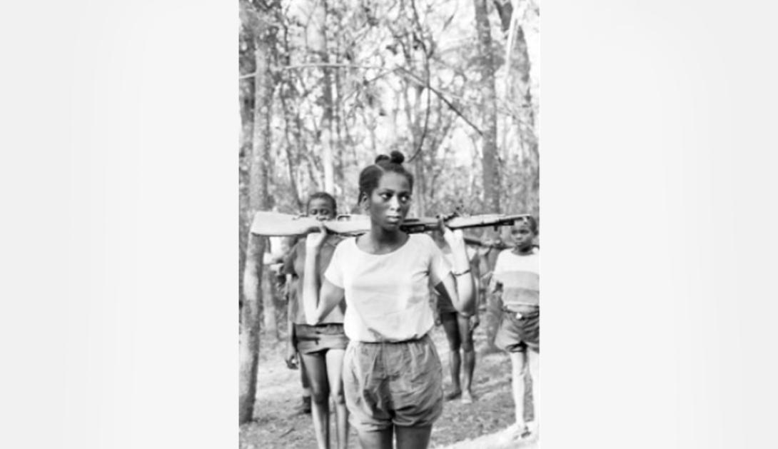 Black-and-white photo of a young woman walking through a wooded area with a rifle across her shoulders, with two youths behind her